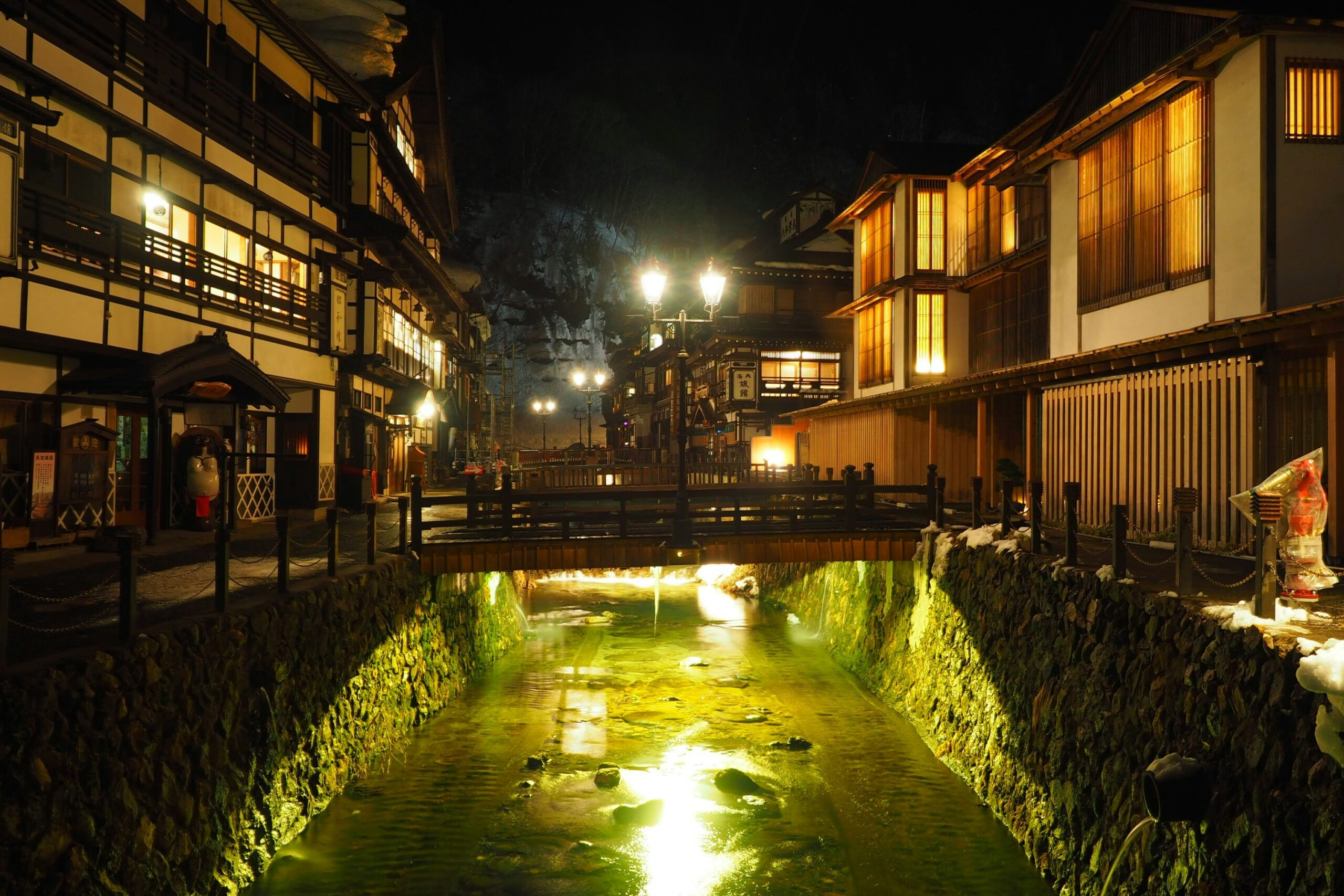 Snowy Ginzan Onsen street in Yamagata with lantern-lit Taisho-era ryokan and steam rising over the river.